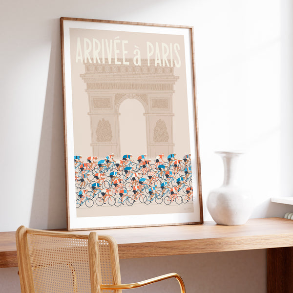 This is a photo of a contemporary style cycling art print in ecru, the poster features an illustrated peloton riding past the Arc de Triomphe in Paris, with the words 'Arrivee a Paris' in a white modern font displayed above the Arc. The print is framed in a plain oak frame and is sat on a wooden desk and leaning against a white wall.