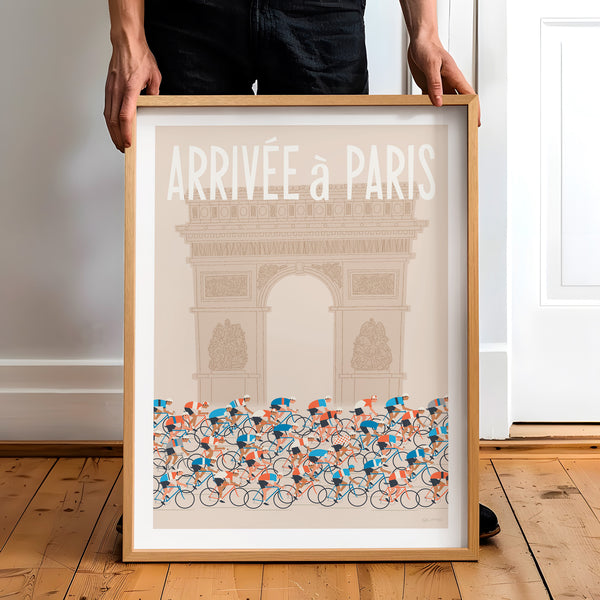 This is a photo of a contemporary style cycling art print in ecru, the framed poster is held by a man and features a peloton riding past the Arc de Triomphe in Paris, with the words 'Arrivee a Paris' in a white modern font displayed above the Arc.