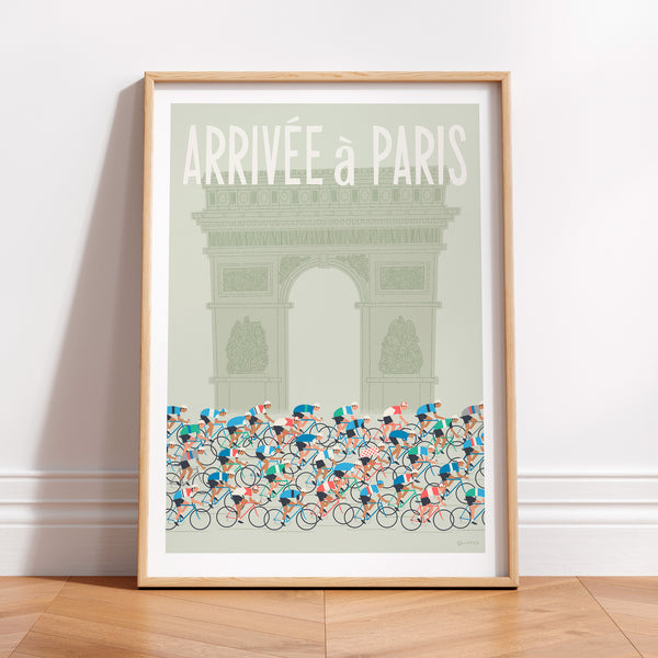 This is a photo of a contemporary style cycling art print in sage green, the poster features a peloton riding past the Arc de Triomphe in Paris, with the words 'Arrivee a Paris' in a white modern font displayed above the Arc. The print is framed in a plain oak frame and is sat on a wooden floor and leaning against a white wall.