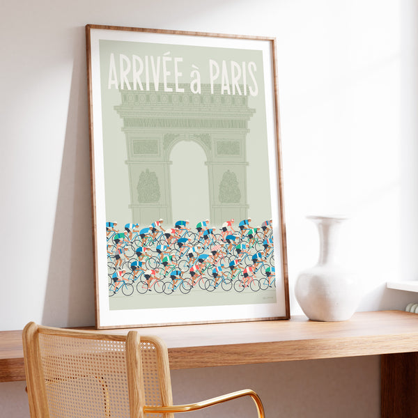This is a photo of a contemporary style cycling art print in sage green, the poster features an illustrated peloton riding past the Arc de Triomphe in Paris, with the words 'Arrivee a Paris' in a white modern font displayed above the Arc. The print is framed in a plain oak frame and is sat on a wooden desk and leaning against a white wall.
