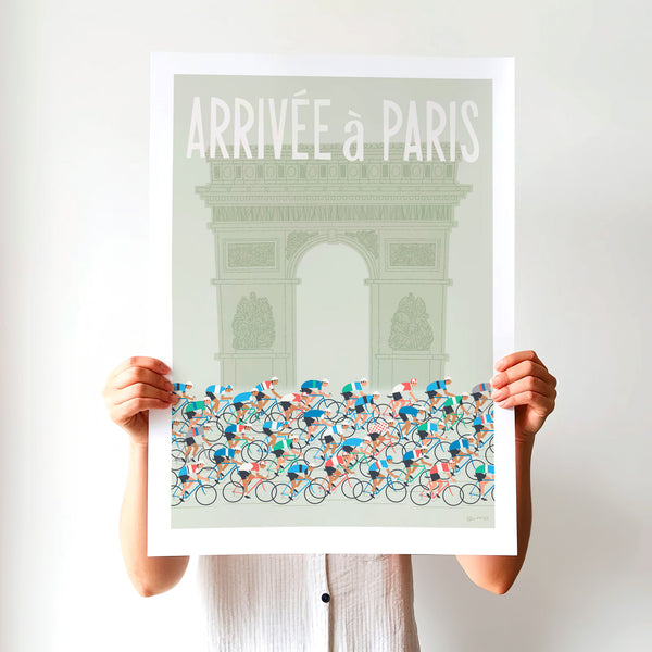 This is a photo of a contemporary style cycling art print in sage green, the unframed poster is held by a woman and features a peloton riding past the Arc de Triomphe in Paris, with the words 'Arrivee a Paris' in a white modern font displayed above the Arc.