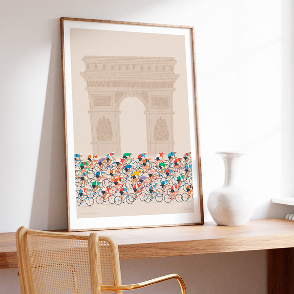 This is a photo of a contemporary style cycling art print in ecru, the poster features a peloton riding past the Arc de Triomphe in Paris. The print is framed in a plain oak frame and is sat on a wooden desk and leaning against a white wall.