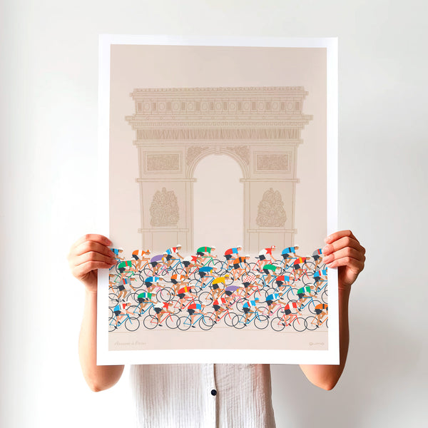 This is a photo of a contemporary cycling art print in ecru, the print features a colourful peloton riding past the Arc de Triomphe in Paris. The unframed print is held up by a woman against a white wall.