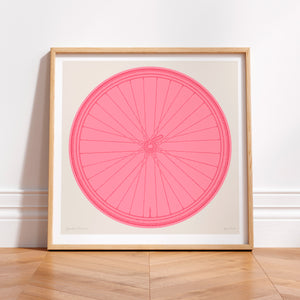 This picture of a contemporary cycling art print features a colourful illustration of a bicycle wheel in giro pink, beneath it in a hand-written font is the phrase: Spokes Person. The print is framed in a simple oak frame and is sat on a wooden herringbone style floor and leaning against a white wall.