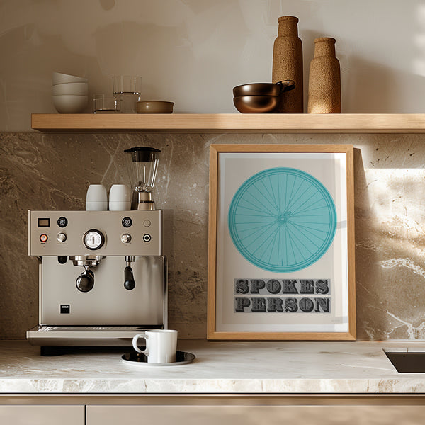 This picture of a contemporary cycling art print features an illustration of a bicycle wheel in coppi blue, beneath it in a bold, decorative font is the phrase: 'Spokes Person'. The print is framed with a light oak frame, sat on a light marble kitchen worktop, next to a stainless steel espresso coffee machine and leaning against a cream wall, with a wooden shelf above.