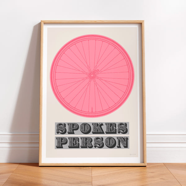 This picture of a contemporary cycling art print features an illustration of a bicycle wheel in giro pink, beneath it in a bold, decorative font is the phrase: 'Spokes Person'. The print is framed with a light oak frame, sat on a wooden floor and leaning against a white wall.