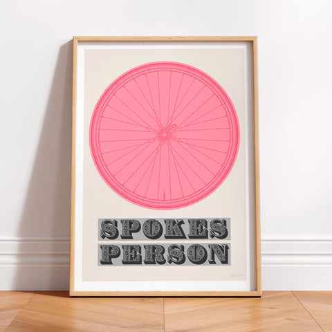 This picture of a contemporary cycling art print features an illustration of a bicycle wheel in giro pink, beneath it in a bold, decorative font is the phrase: 'Spokes Person'. The print is framed with a light oak frame, sat on a wooden floor and leaning against a white wall.