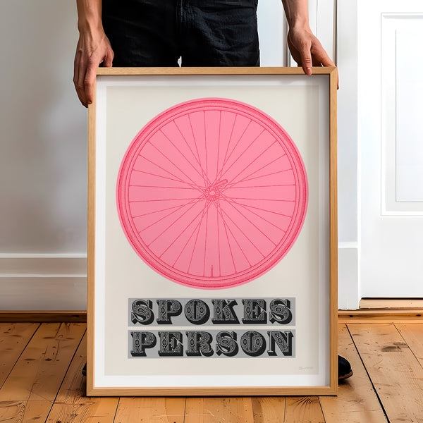 This picture of a contemporary cycling art print features an illustration of a bicycle wheel in giro pink, beneath it in a bold, decorative font is the phrase: 'Spokes Person'. The framed print is held by a man.