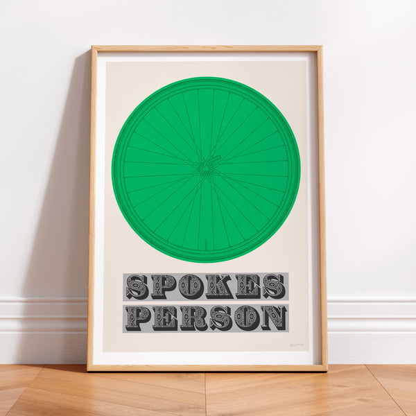 This picture of a contemporary cycling art print features an illustration of a bicycle wheel in kelly green, beneath it in a bold, decorative font is the phrase: 'Spokes Person'. The print is framed with a light oak frame, sat on a wooden floor and leaning against a white wall.