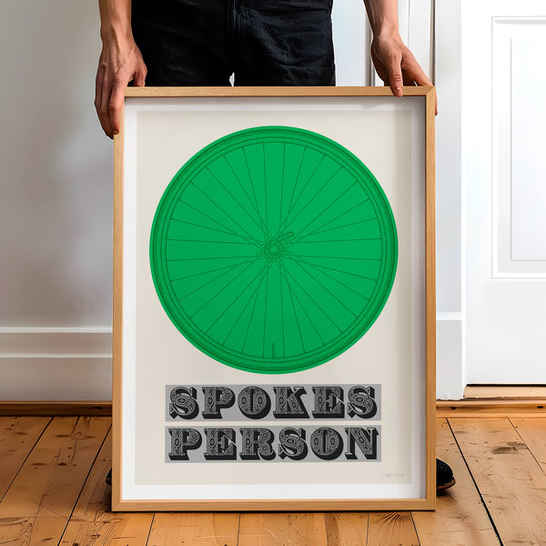 This picture of a contemporary cycling art print features an illustration of a bicycle wheel in kelly green, beneath it in a bold, decorative font is the phrase: 'Spokes Person'. The framed print is held by a man.