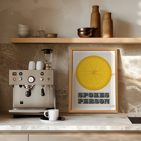 This picture of a contemporary cycling art print features an illustration of a bicycle wheel in tour yellow, beneath it in a bold, decorative font is the phrase: 'Spokes Person'. The print is framed with a light oak frame, sat on a light marble kitchen worktop, next to a stainless steel espresso coffee machine and leaning against a cream wall, with a wooden shelf above.