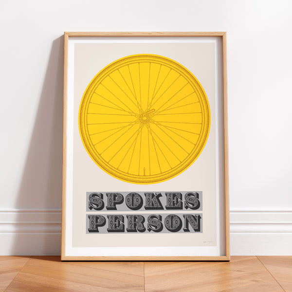 This picture of a contemporary cycling art print features an illustration of a bicycle wheel in tour yellow, beneath it in a bold, decorative font is the phrase: 'Spokes Person'. The print is framed with a light oak frame, sat on a wooden floor and leaning against a white wall.