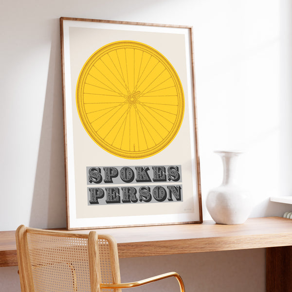 This picture of a contemporary cycling art print features an illustration of a bicycle wheel in tour yellow, beneath it in a bold, decorative font is the phrase: 'Spokes Person'. The framed print is sat on a modern style wooden desk