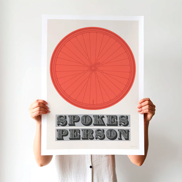 This picture of a contemporary cycling art print features an illustration of a bicycle wheel in vuelta red, beneath it in a bold, decorative font is the phrase: 'Spokes Person'. The unframed print is held up by a girl.