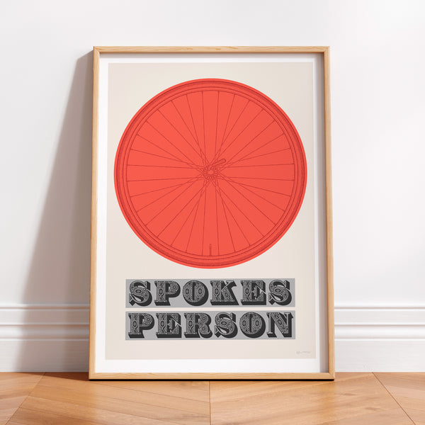 This picture of a contemporary cycling art print features an illustration of a bicycle wheel in vuelta red, beneath it in a bold, decorative font is the phrase: 'Spokes Person'. The print is framed with a light oak frame, sat on a wooden floor and leaning against a white wall.