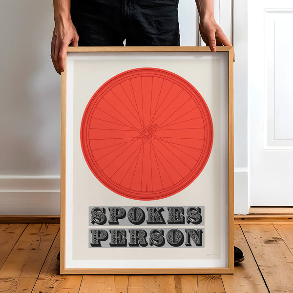 This picture of a contemporary cycling art print features an illustration of a bicycle wheel in vuelta red, beneath it in a bold, decorative font is the phrase: 'Spokes Person'. The framed print is held by a man.