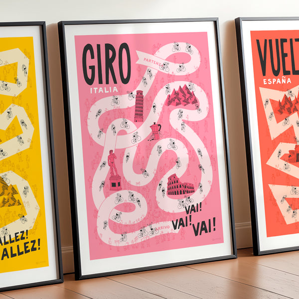 Set of three Grand Tour cycling posters—Giro, Tour, and Vuelta, focussing on the Giro design in the centre and with the Tour and Vuelta designs shown cropped on either side. The print is framed and leaning against a wall and features hand drawn illustrations of a pale pink road against a darker pink background, including riders, spectators and famous Italian landmarks.
