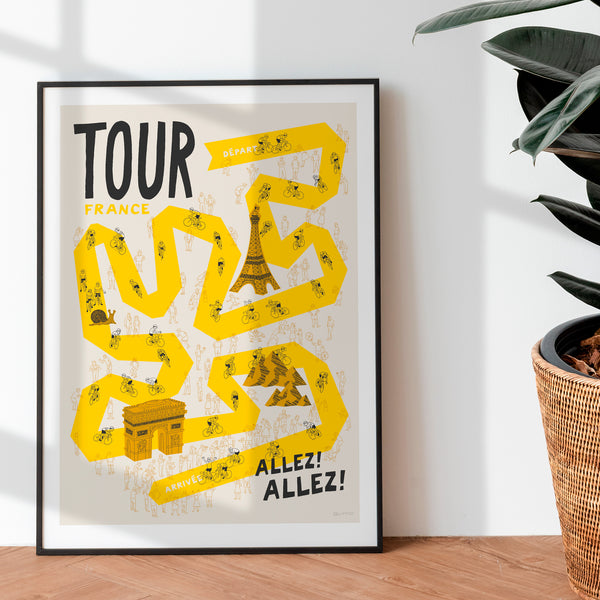 Framed poster with 'Tour de France' design propped up against a wall with a plant.