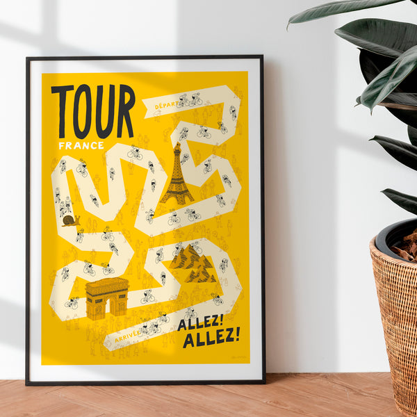 Grand Tour cycling print, featuring a pale yellow road on a darker yellow background, with hand drawn illustrations of cyclists, spectators and famous Fench landmarks. It is displayed in a black frame leaning against a white wall, with a rubber plant nearby on a wooden floor.