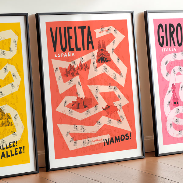 Set of three Grand Tour cycling posters—Giro, Tour, and Vuelta, focussing on the Vuelta design in the centre and with the Tour and Giro designs shown cropped on either side. The print is framed and leaning against a wall and features hand drawn illustrations of a pale red road against a darker red background, along with cyclists, spectators and famous Spanish landmarks.