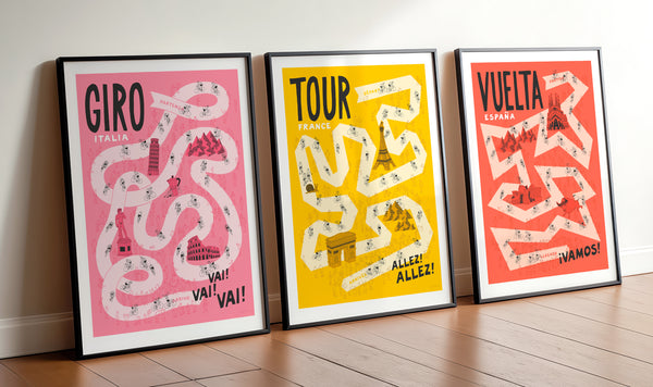 Grand Tour cycling art poster set, featuring Giro, Tour, and Vuelta designs, with pale pink, yellow, and red roads, featuring hand drawn illustrations of cyclists, fans and famous landmarks. The designs of each are set against darker pink, yellow and red backgrounds respectively. The prints are framed in black and displayed side by side and propped on a wooden floor, leaning against a wall.
