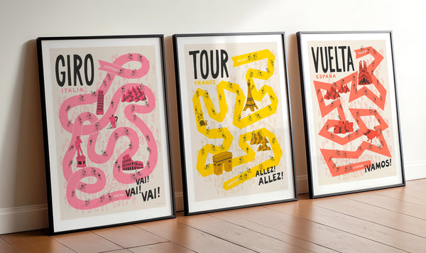 Three framed posters with 'Giro', 'Tour', and 'Vuelta' designs on a wooden floor.
