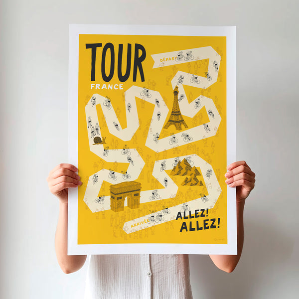Unframed Tour Grand Tour cycling poster being held up, featuring a pale yellow winding road on a darker yellow background, including hand drawn illustrations of cyclists, fans and a selection of famous French landmarks.