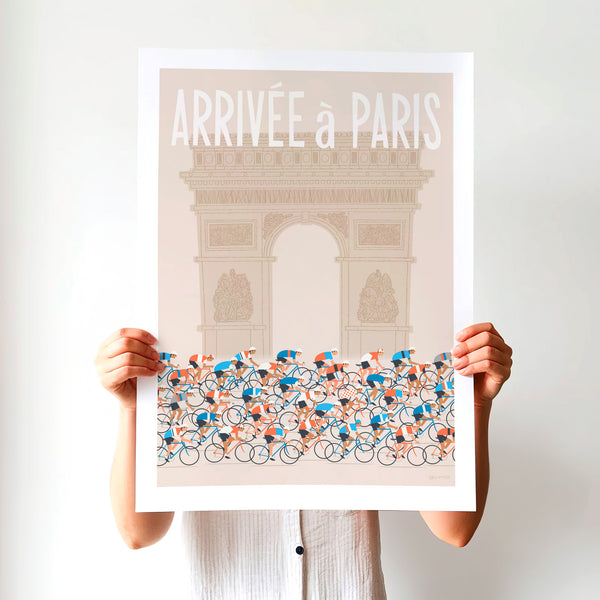 This is a photo of a contemporary style cycling art print in ecru, the unframed poster is held by a woman and features a peloton riding past the Arc de Triomphe in Paris, with the words 'Arrivee a Paris' in a white modern font displayed above the Arc.  
