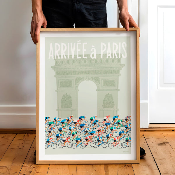 This is a photo of a contemporary style cycling art print in sage green, the framed poster is held by a man and features a peloton riding past the Arc de Triomphe in Paris, with the words 'Arrivee a Paris' in a white modern font displayed above the Arc. 