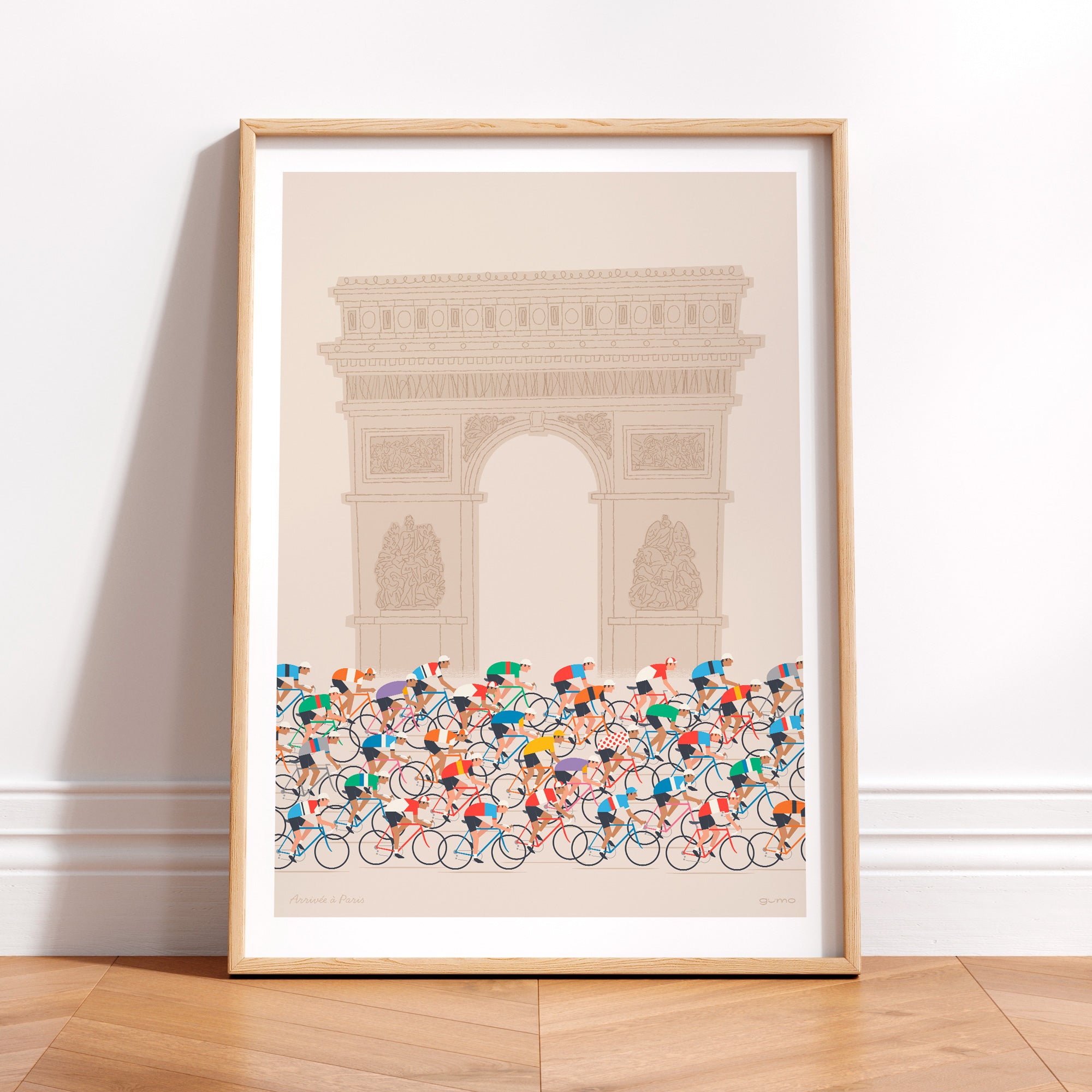 This is a photo of a contemporary cycling art print in ecru, it features the peloton riding past the Arc de Triomphe in Paris. The print is framed in a simple oak frame and is sat on a wooden herringbone style floor and leaning against a white wall.